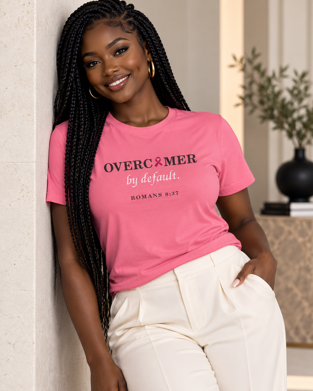 A woman with long black braids leans against a light beige wall, wearing a pink premium T-shirt with the text 'OVERCOMER by default. ROMANS 8:37' by Press & Amen, and cream-colored pants.