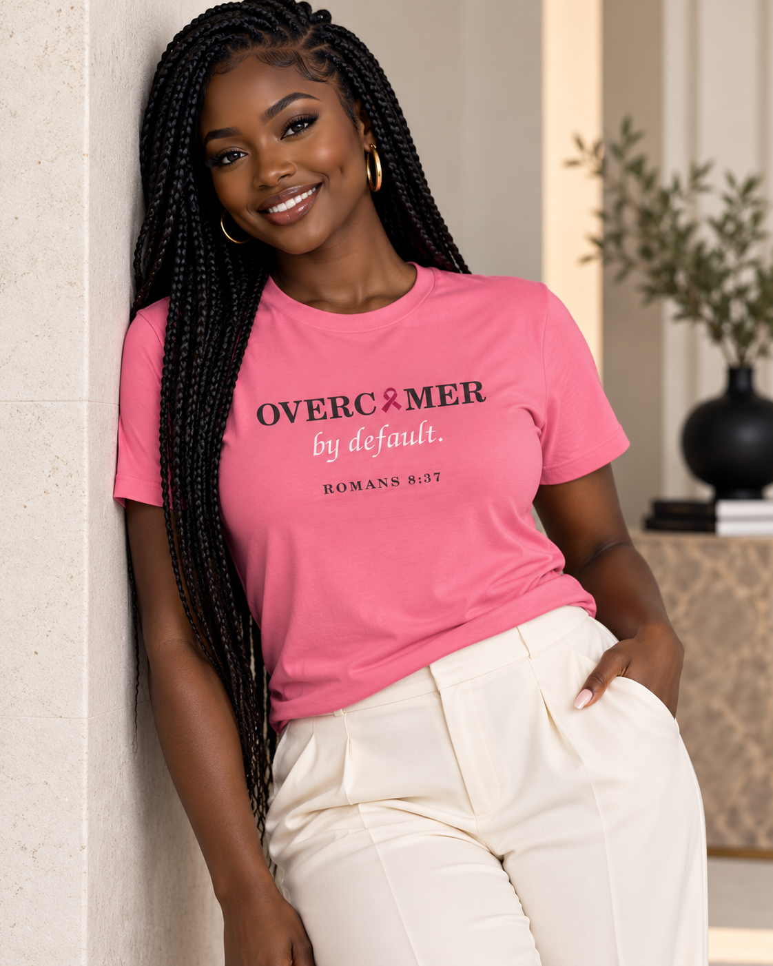 A woman with long black braids leans against a light beige wall, wearing a pink premium T-shirt with the text 'OVERCOMER by default. ROMANS 8:37' by Press & Amen, and cream-colored pants.